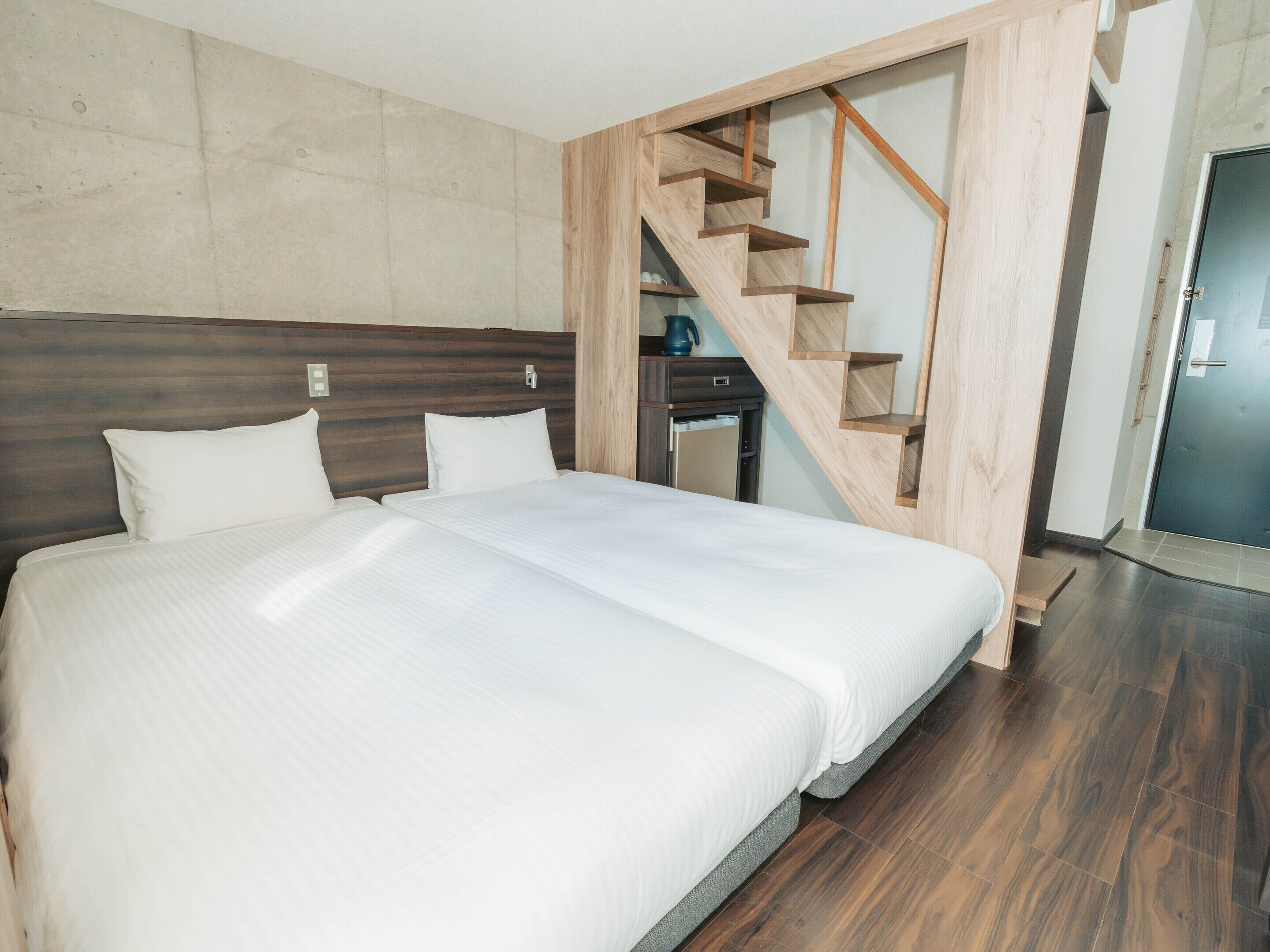 A modern hotel room with a large bed, a wooden staircase, and a kitchenette.