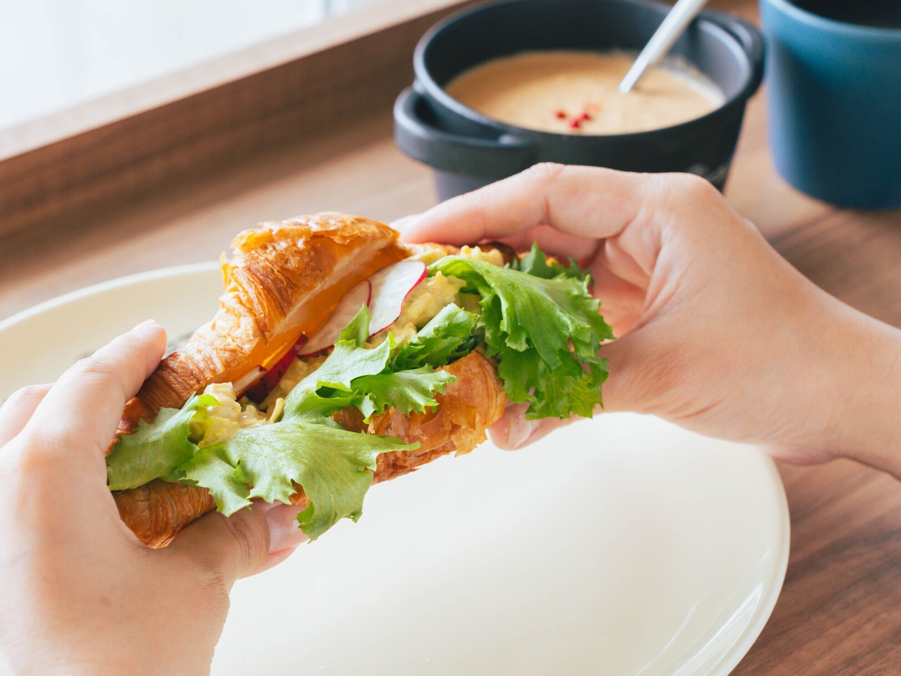A person holding a sandwich with visible lettuce and a folded bread, a bowl of soup, and a cup on a table.