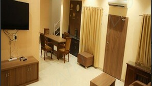 A living room with a sofa, coffee table, and TV. There is a dining area with chairs and a table. A small cabinet is visible, and a door is in the background. - SkyWater Villas And Lifestyle (Igatpuri)