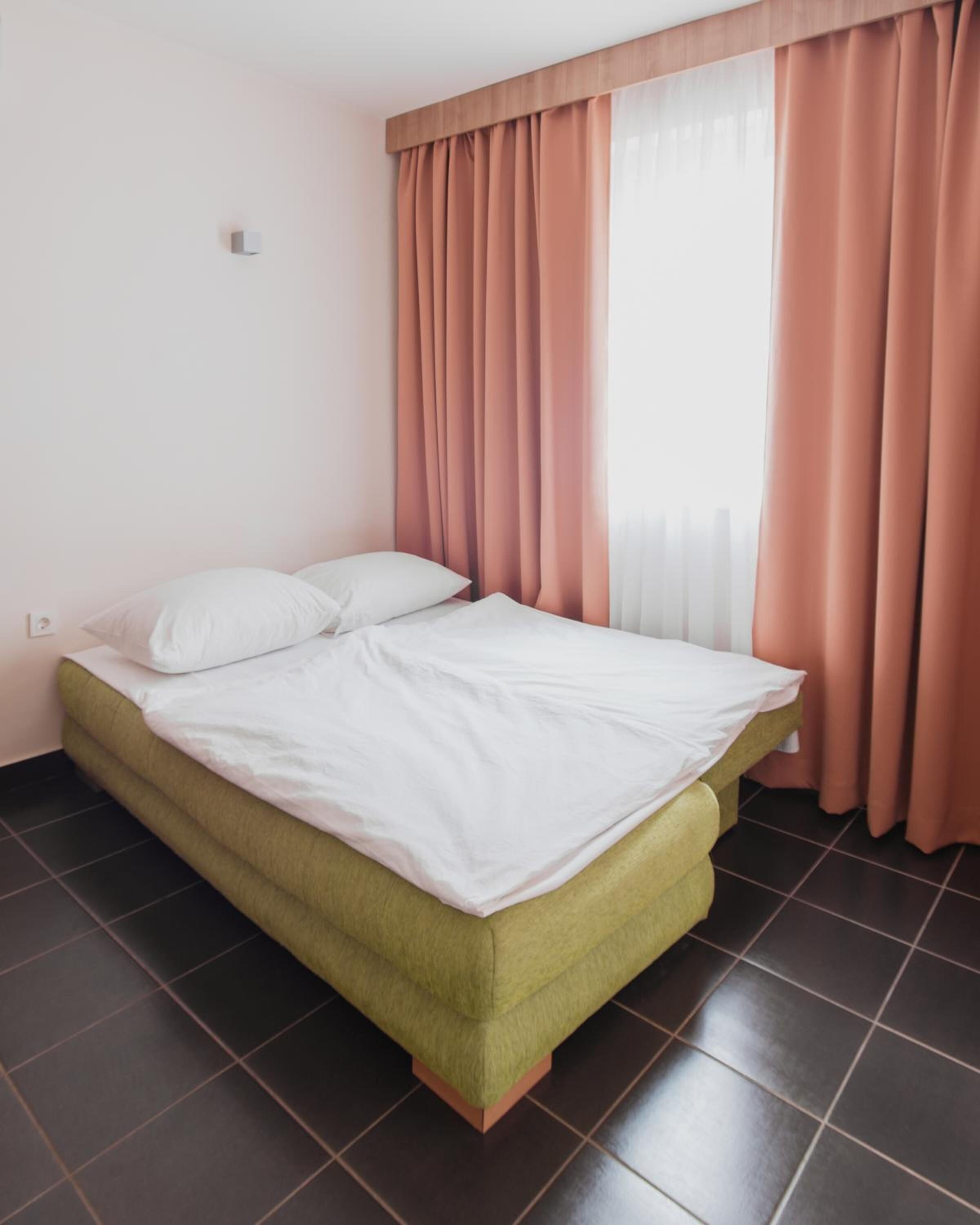 A single bed with a green upholstered headboard in a room with dark tiled flooring and pink curtains.