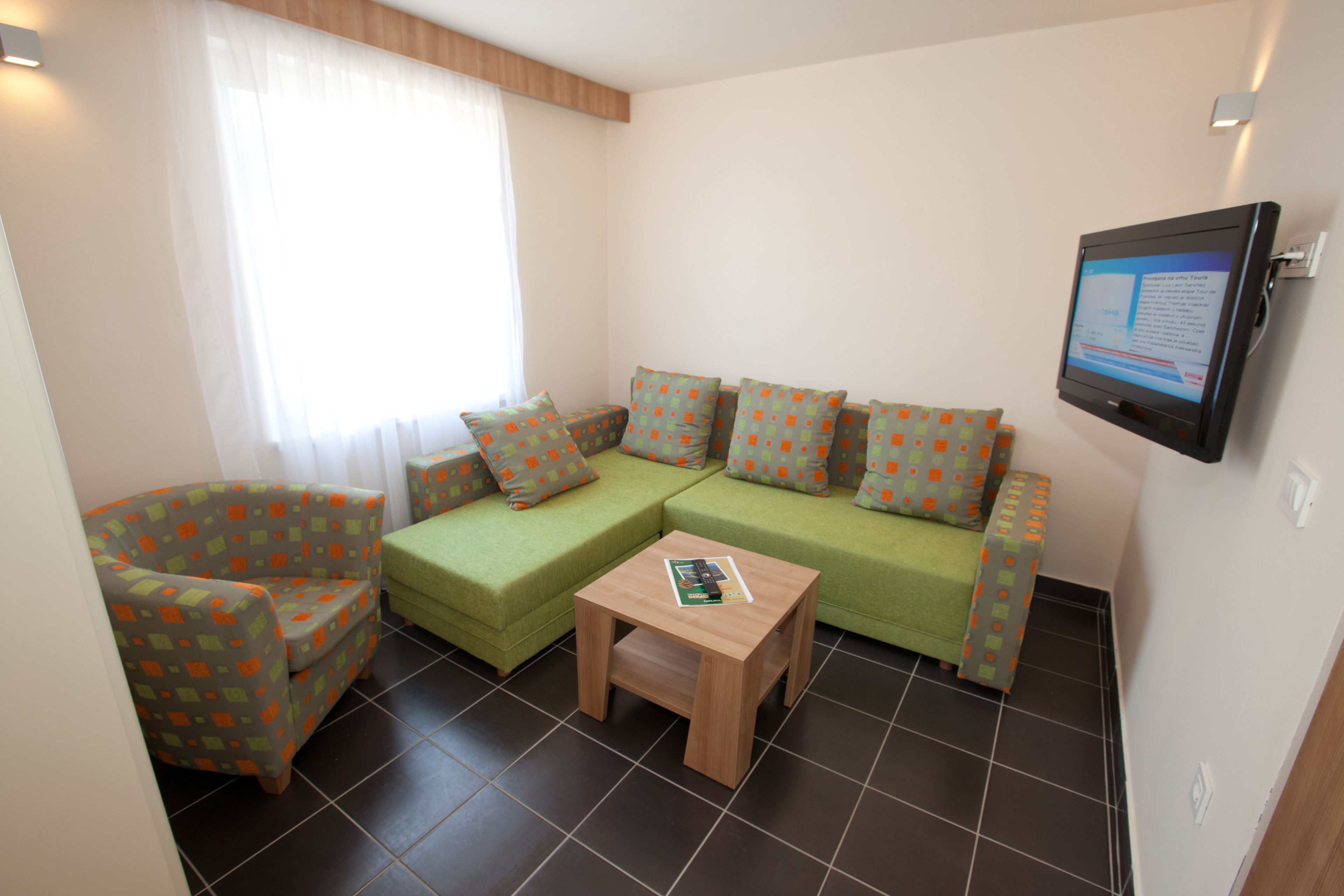 Un petit salon moderne avec un canapé vert, un fauteuil assorti et une table basse en bois. Un téléviseur à écran plat est fixé au mur.