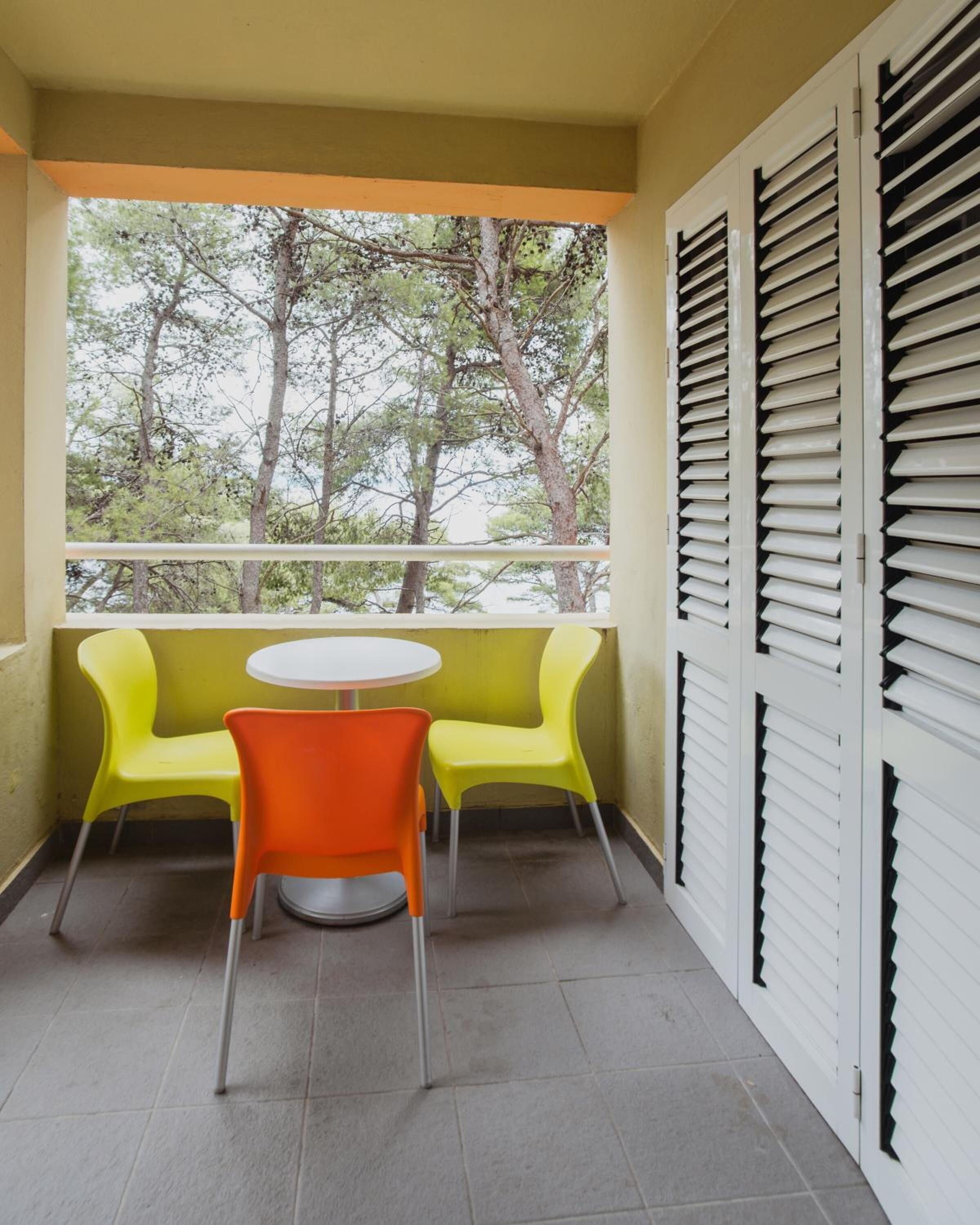 A balcony with a round table and two chairs, one orange and one yellow, with a view of trees through the window.