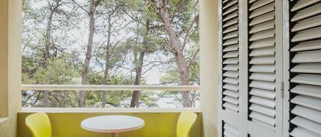 A balcony with a round table and two chairs, one orange and one yellow, with a view of trees through the window.