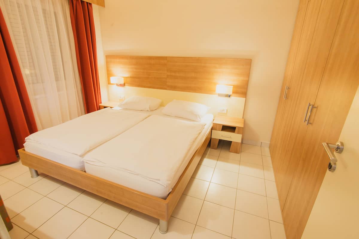 Hotellihuone, jossa on puinen sänky, kaksi yöpöytää valaisimineen, puinen vaatekaappi ja laattalattia.