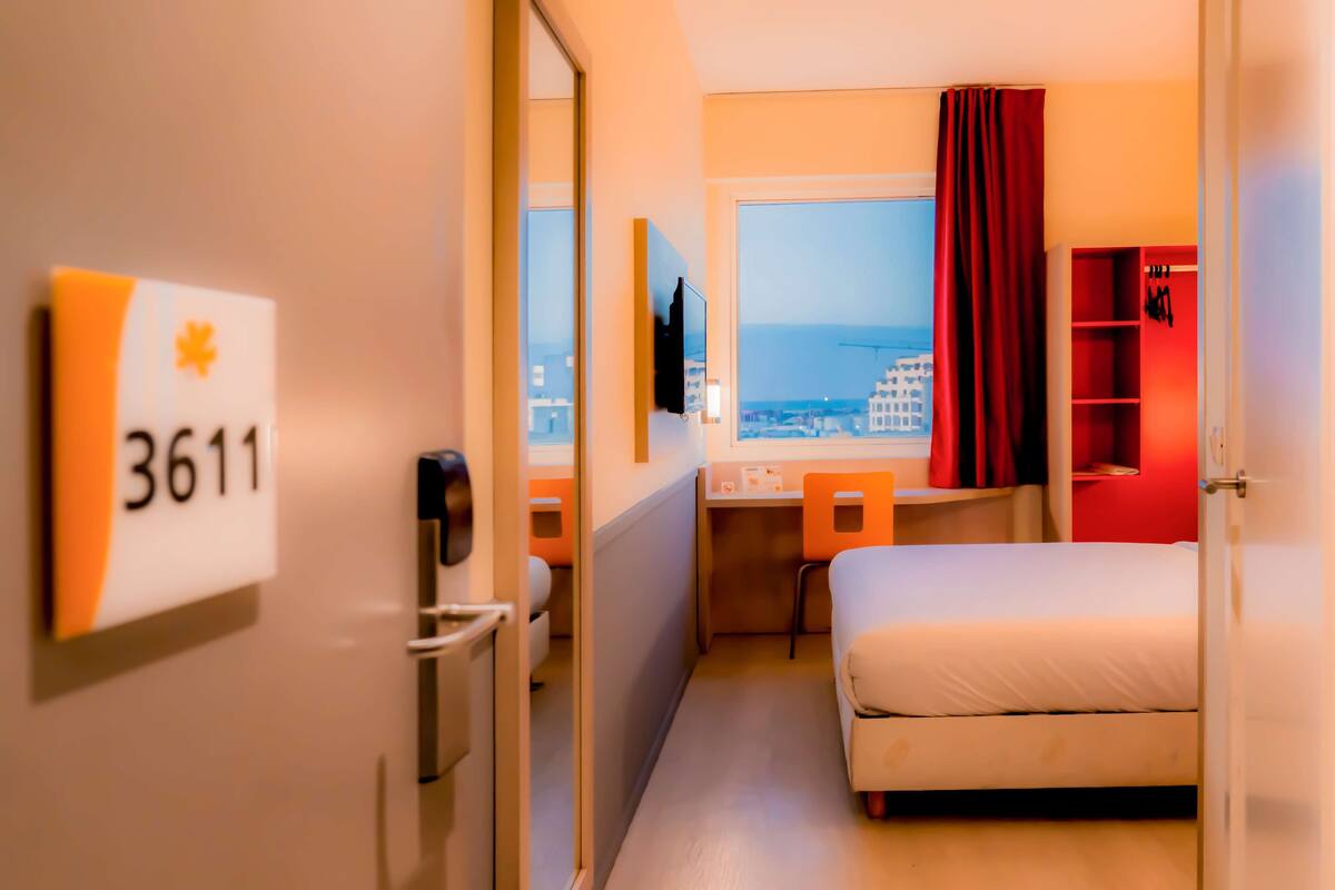 A hotel room with a bed, a desk, a chair, a TV, a window with a view of the city, and a red curtain.