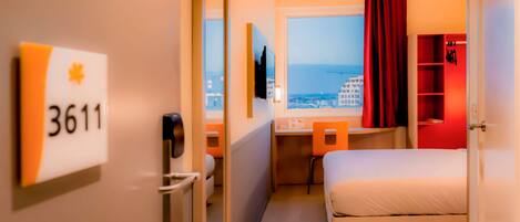 A hotel room with a bed, a desk, a chair, a TV, a window with a view of the city, and a red curtain.
