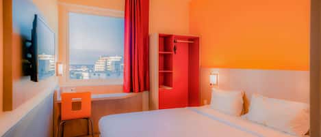 Una habitación de hotel con una cama, un escritorio, una silla, un armario rojo y una ventana con vistas a edificios.