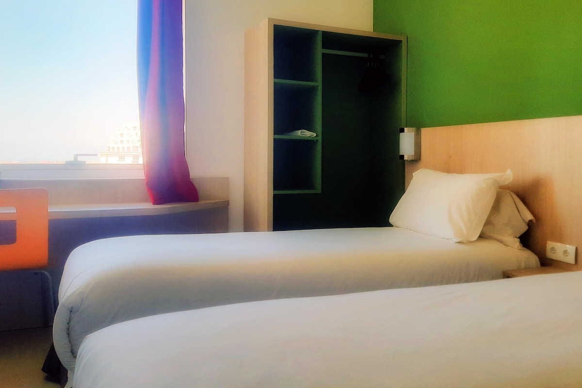 A hotel room with two beds, a desk, and a window with a view of buildings.