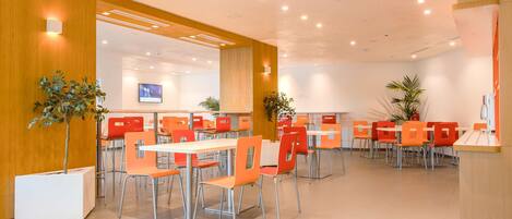 A modern dining area with orange chairs and white tables, wooden paneling on the walls, and ceiling lights.