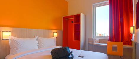 A hotel room with a bed, a desk, a chair, a wardrobe, and a window with red curtains.