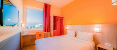 Habitación de hotel con una cama grande, un escritorio, una silla, un televisor, un ventanal con vistas a edificios y un armario rojo.