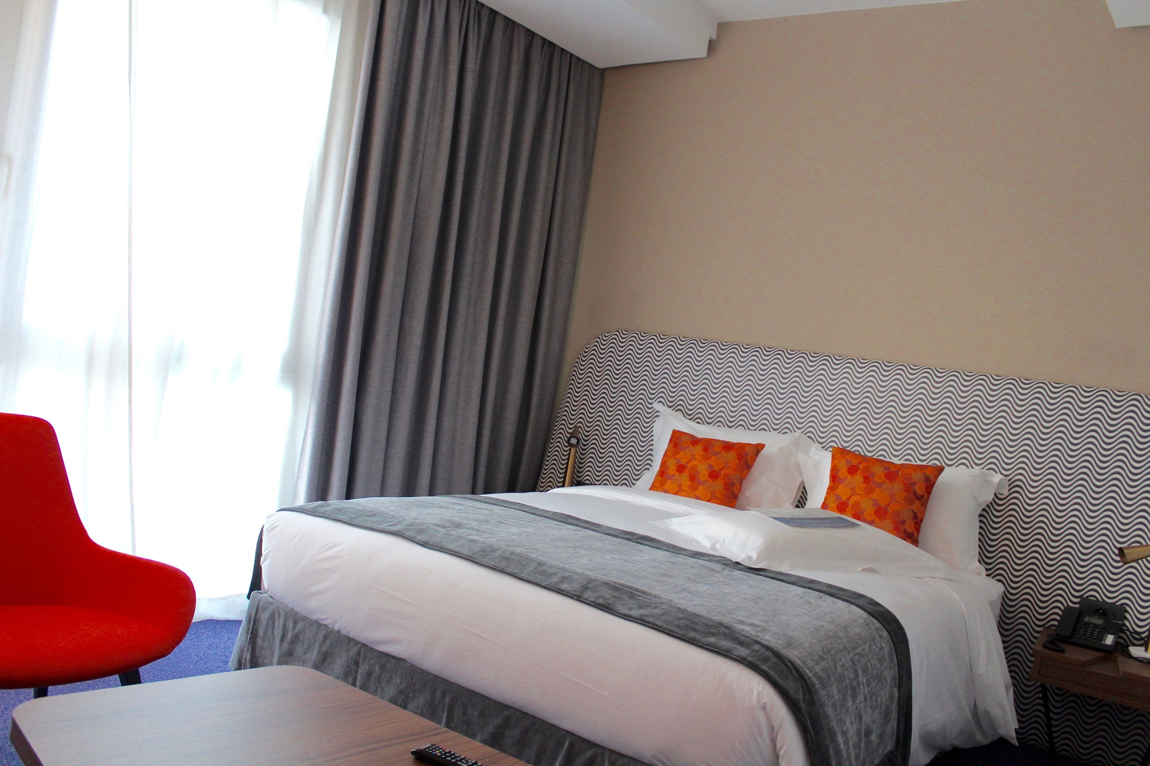 Una habitación de hotel con una cama, una silla roja, una mesita pequeña y una mesita de noche con un teléfono.