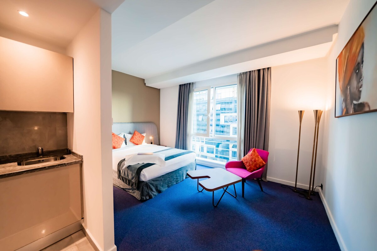A hotel room with a bed, a desk, a chair, a window with curtains, and a painting on the wall.