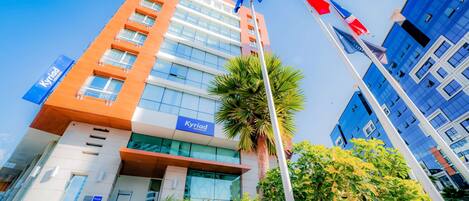 Un moderno edificio de hotel con un letrero destacado de 'Kyriad', varios pisos y un asta con banderas.