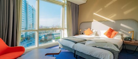 A hotel room with a large window, a bed with multiple pillows, a bedside table with a lamp, and a red chair.