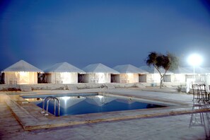 Una piscina con escalera, rodeada de tiendas blancas con techos cónicos, y algunas sillas y mesas cerca.