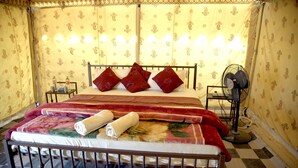 Una cama bien hecha con una colcha roja y blanca, dos almohadas rojas y una toalla blanca enrollada. Hay una mesita de noche pequeña con una lámpara y una botella. La habitación tiene papel tapiz estampado y un suelo a cuadros.