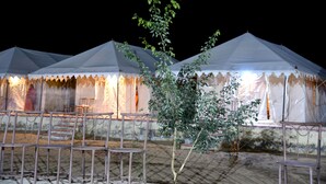 Una hilera de tiendas blancas con sillas metálicas al frente, un pequeño árbol en el centro y un suelo de baldosas.