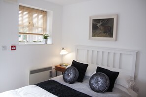 A neatly arranged bedroom with a bed, a bedside table with a lamp, a window with blinds, and a framed picture on the wall. - The Dukes (Leighton Buzzard)