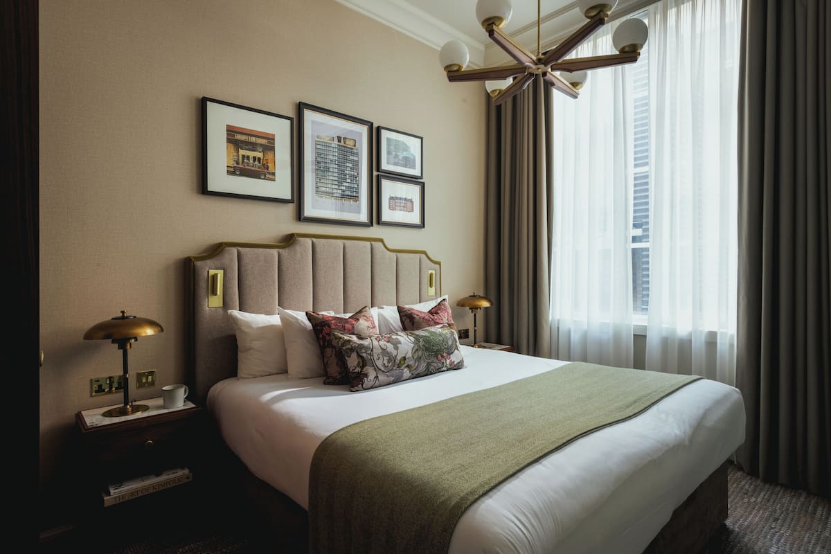 Quarto de hotel com uma cama grande, mesa de cabeceira com abajur, quadros nas paredes e vista para a cidade pela janela.