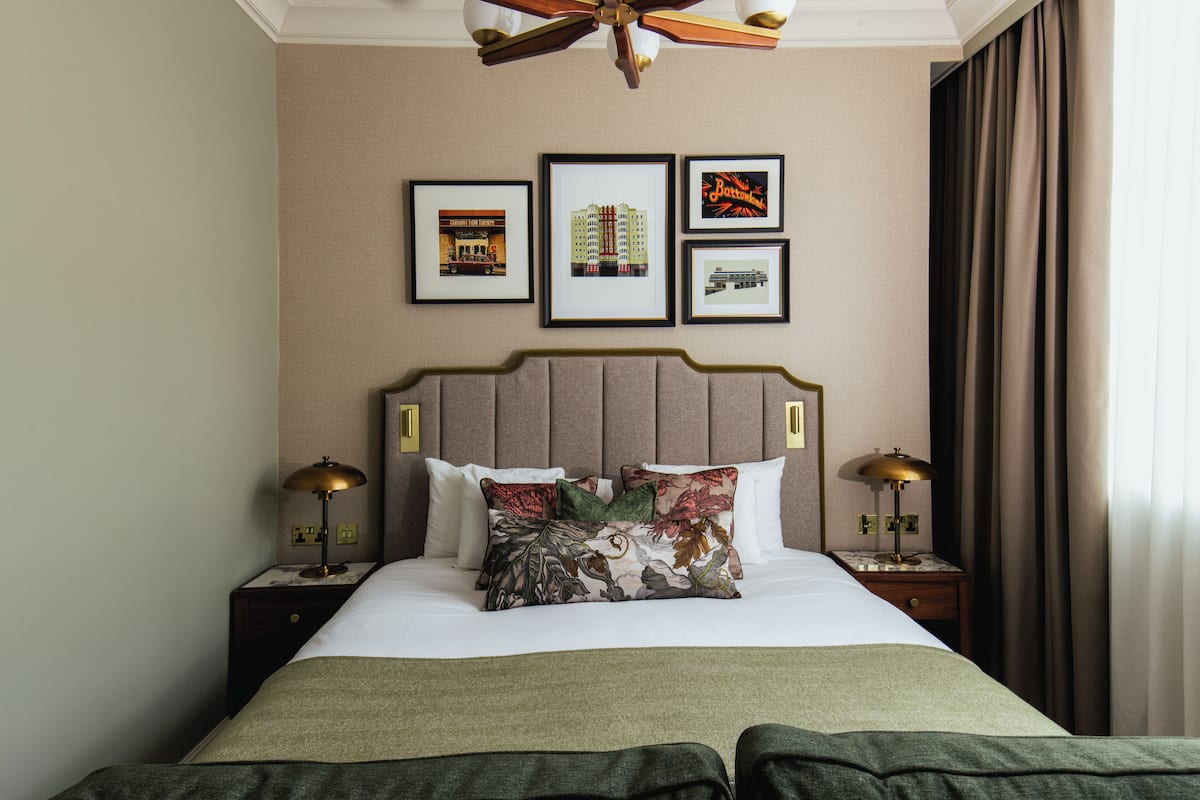 Quarto de hotel com cama, cabeceira, mesas de cabeceira, abajures e quadros nas paredes.