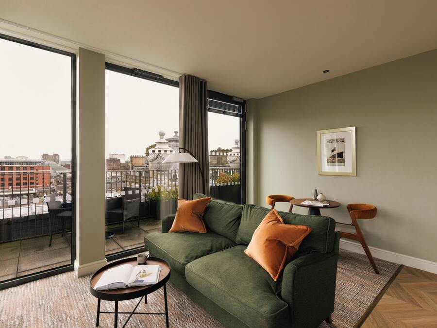 A modern living room with a green sofa, two orange cushions, a small round table, and a chair. There is a large window with a view of buildings and a balcony.