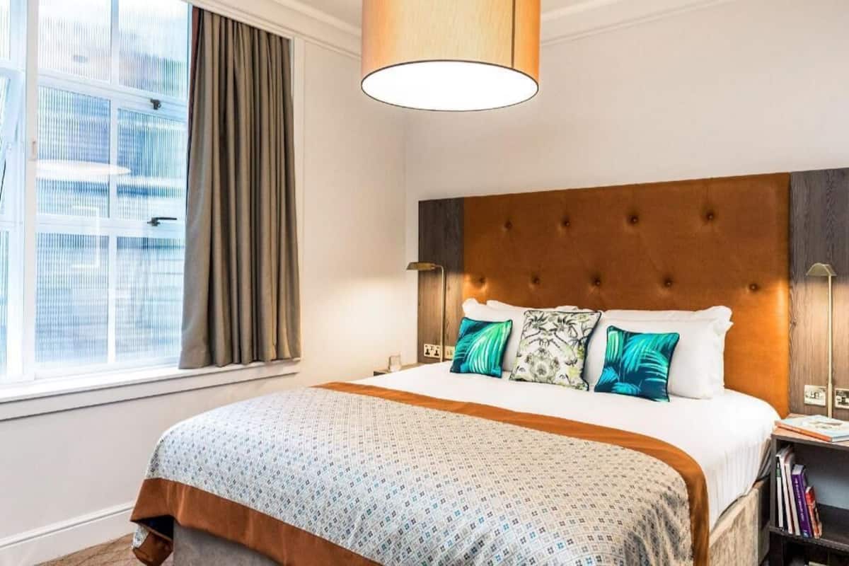 Quarto de hotel com uma cama grande, janela com cortinas, cabeceira e mesa de cabeceira com livros.