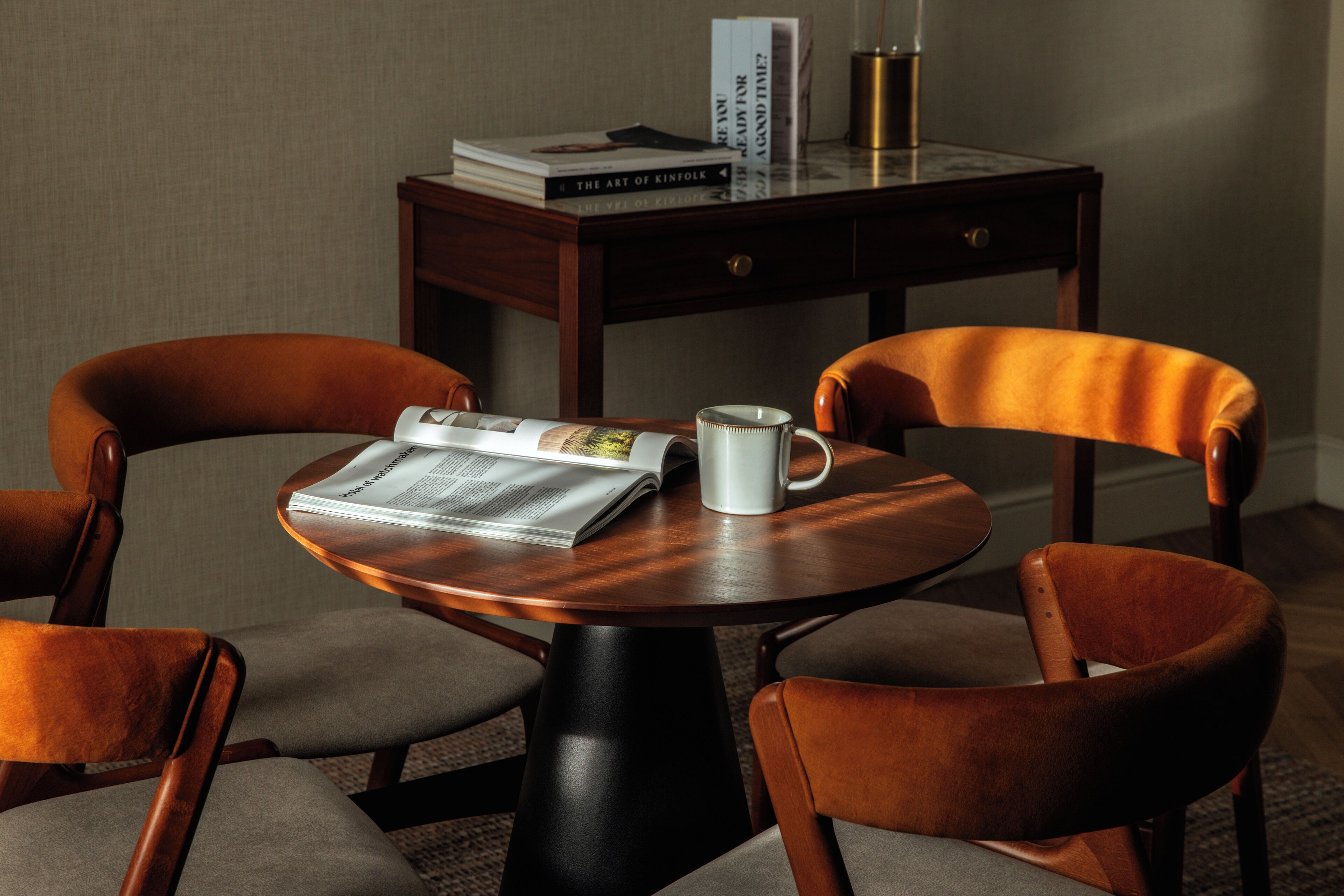A round wooden table with a newspaper, a white mug, and books. There are two brown upholstered chairs around the table.