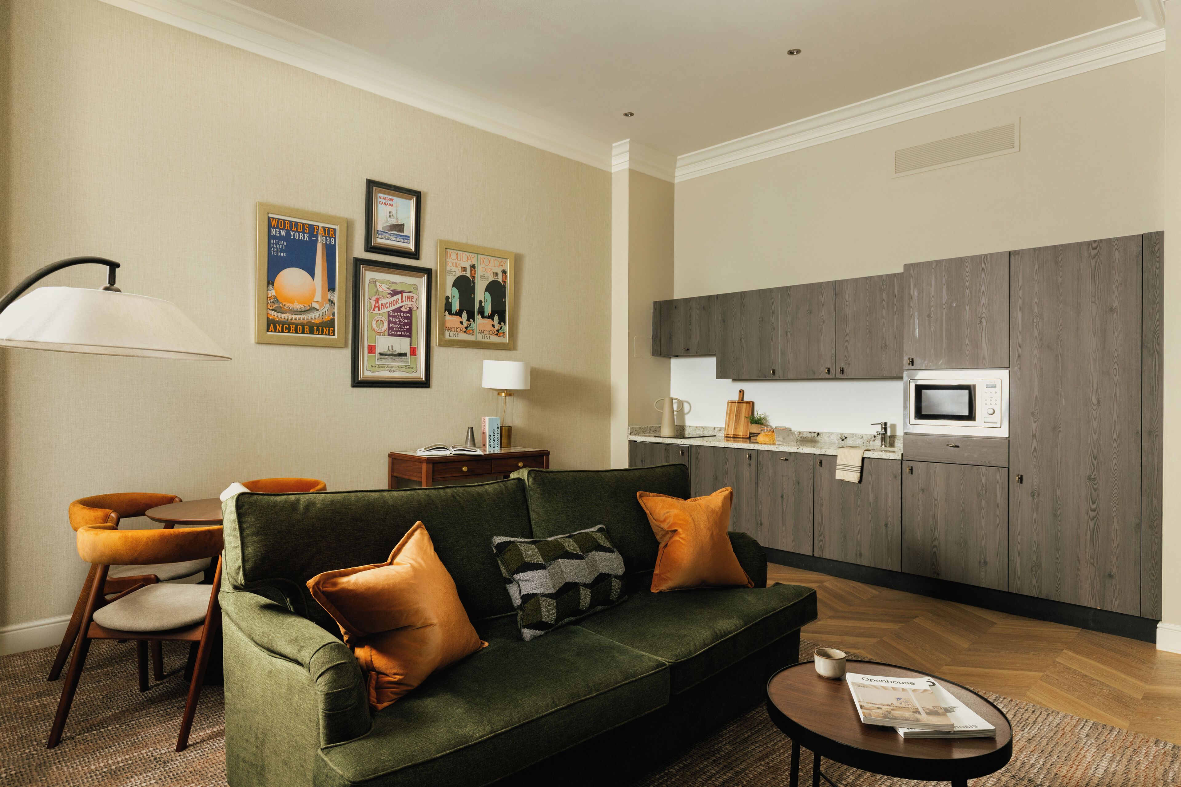 A living room with a green sofa, orange cushions, a coffee table, and a lamp. There is a kitchen area with wooden cabinets and a microwave.