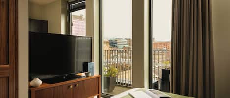 A hotel room with a large flat-screen TV on a wooden cabinet, a bed with a green throw, and a view of the city through large windows.