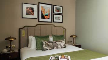 A neatly made bed with green and floral pillows, a green throw blanket, and a magazine on the bed. There are framed pictures on the wall above the bed.
