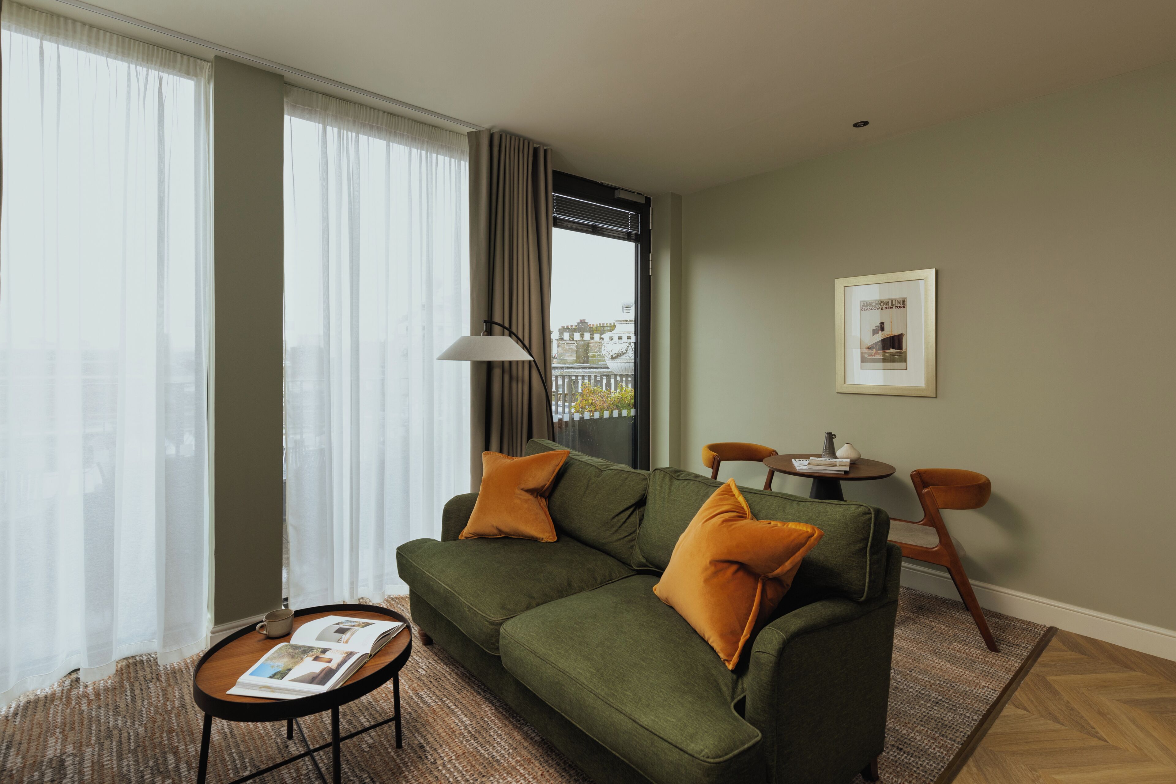 Ein modernes Wohnzimmer mit einem grünen Sofa, zwei orangefarbenen Kissen, einem kleinen runden Tisch mit einer Zeitschrift und einer Lampe.
