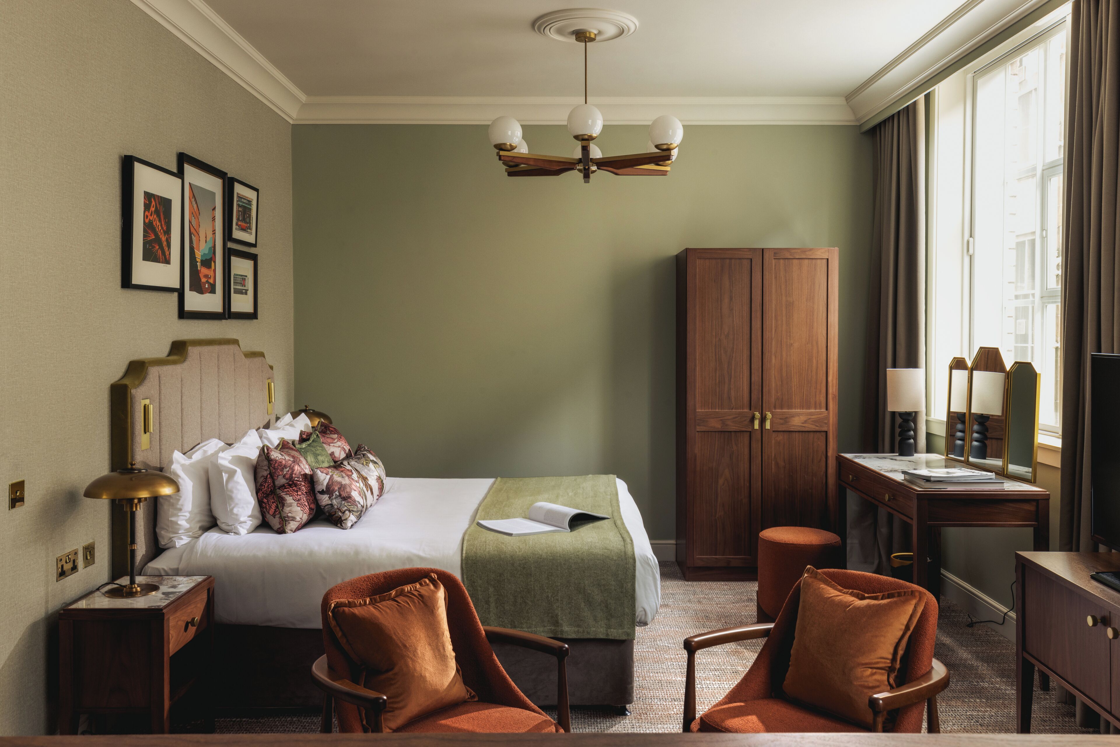 A hotel room with a bed, two chairs, a desk, a wardrobe, and a large window.