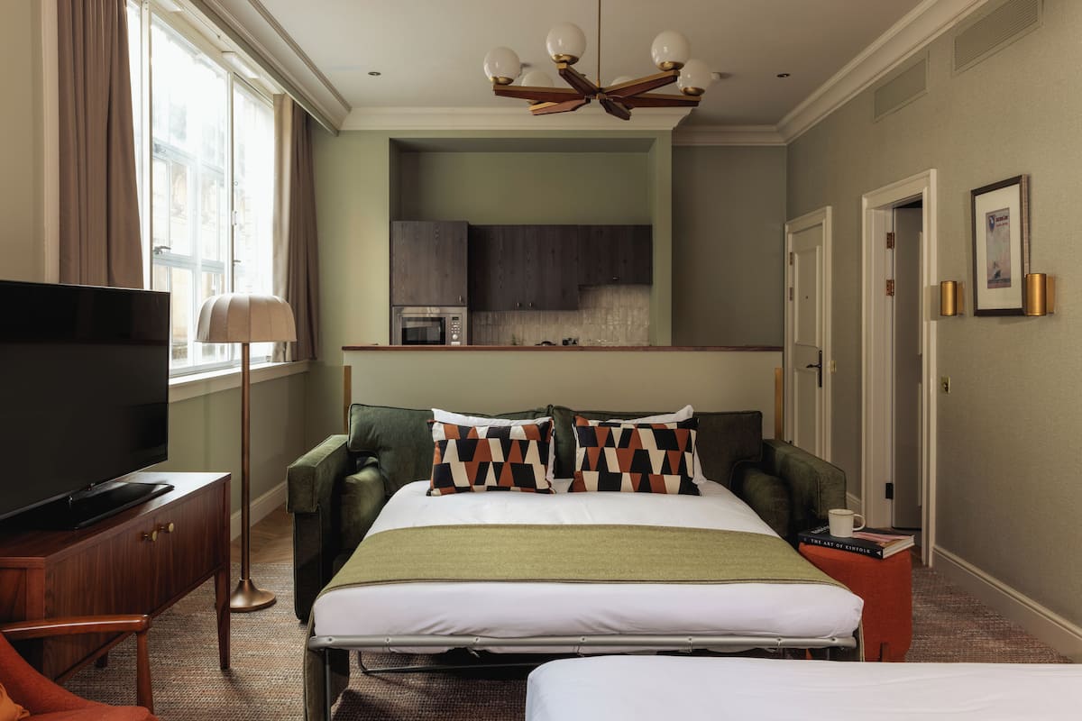 Quarto de hotel com cama, escrivaninha com televisão, cadeira, mesa de cabeceira, luminária, janela com cortinas, cozinha compacta e luminária no teto.