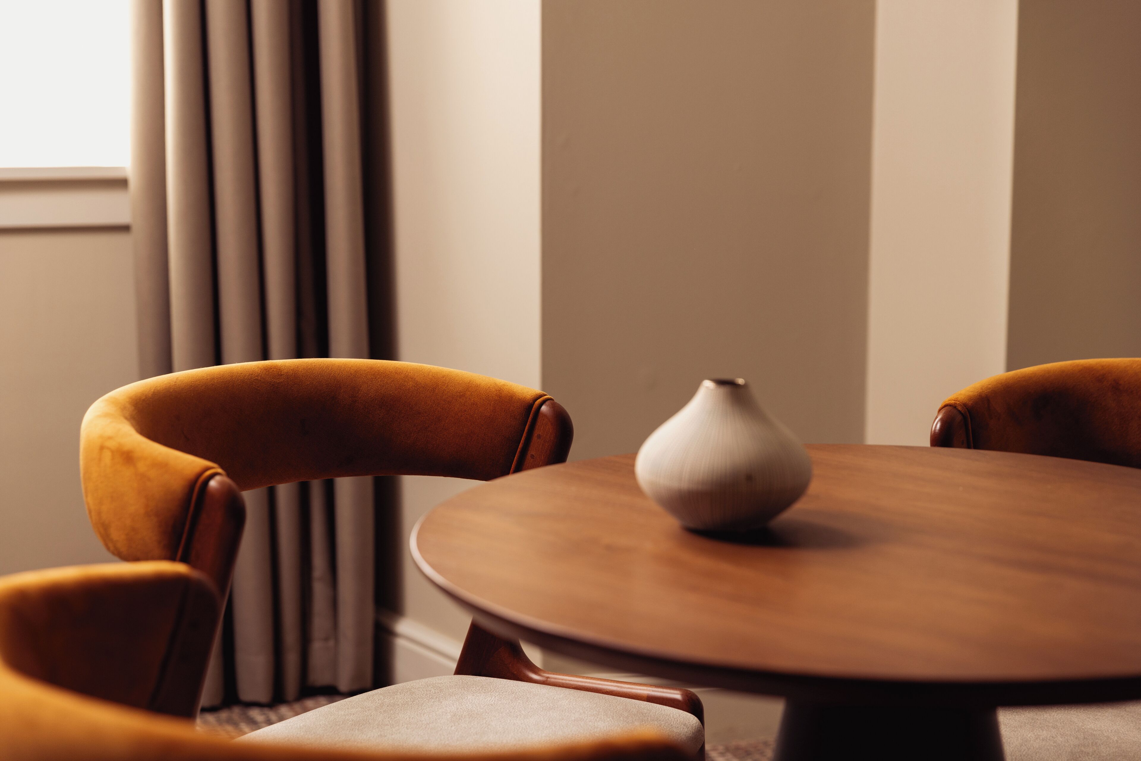 A round wooden table with a white vase in the center, surrounded by four brown upholstered chairs.