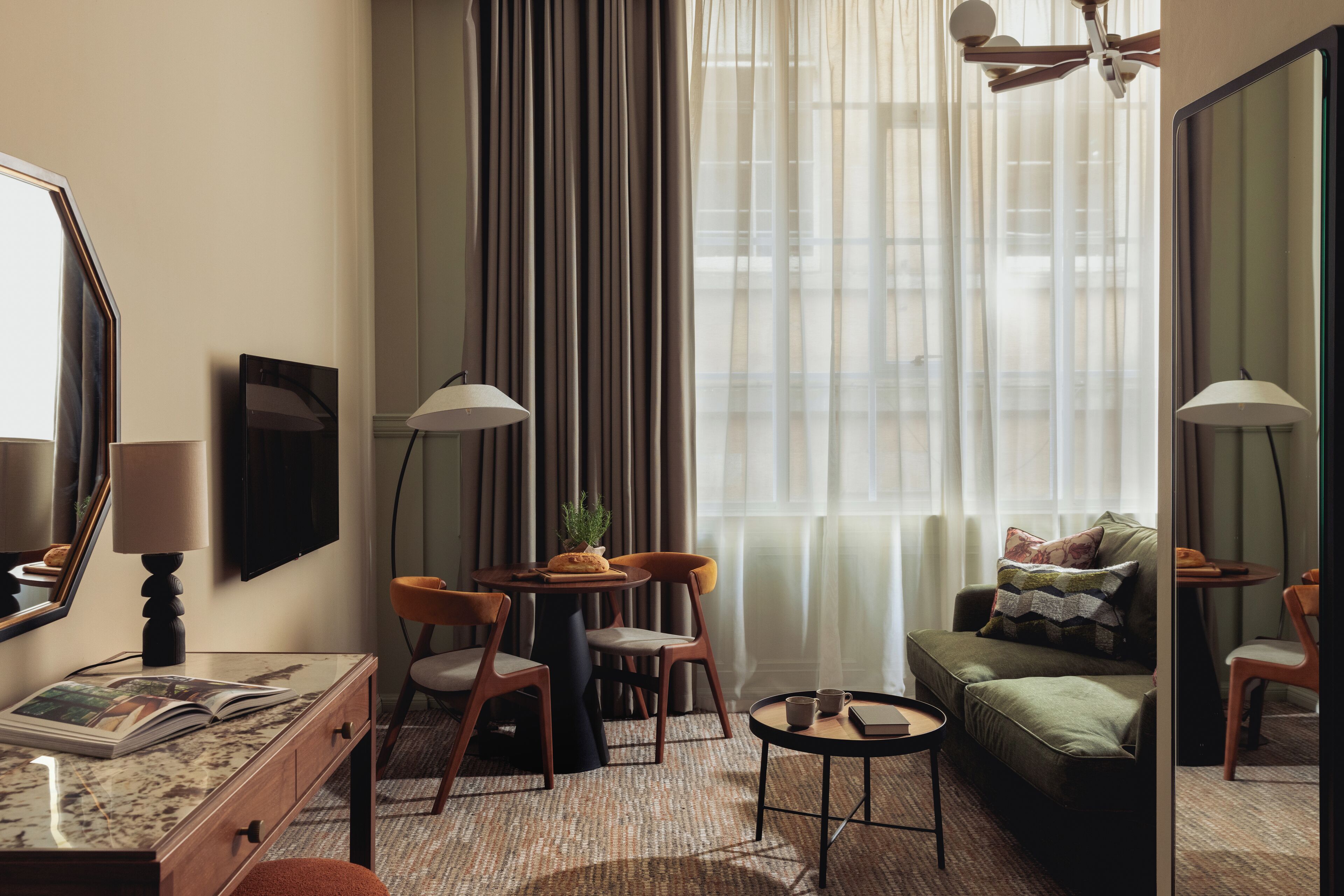 Une chambre d’hôtel comprenant un coin repas, un canapé, un bureau avec une lampe et une grande fenêtre avec des rideaux.