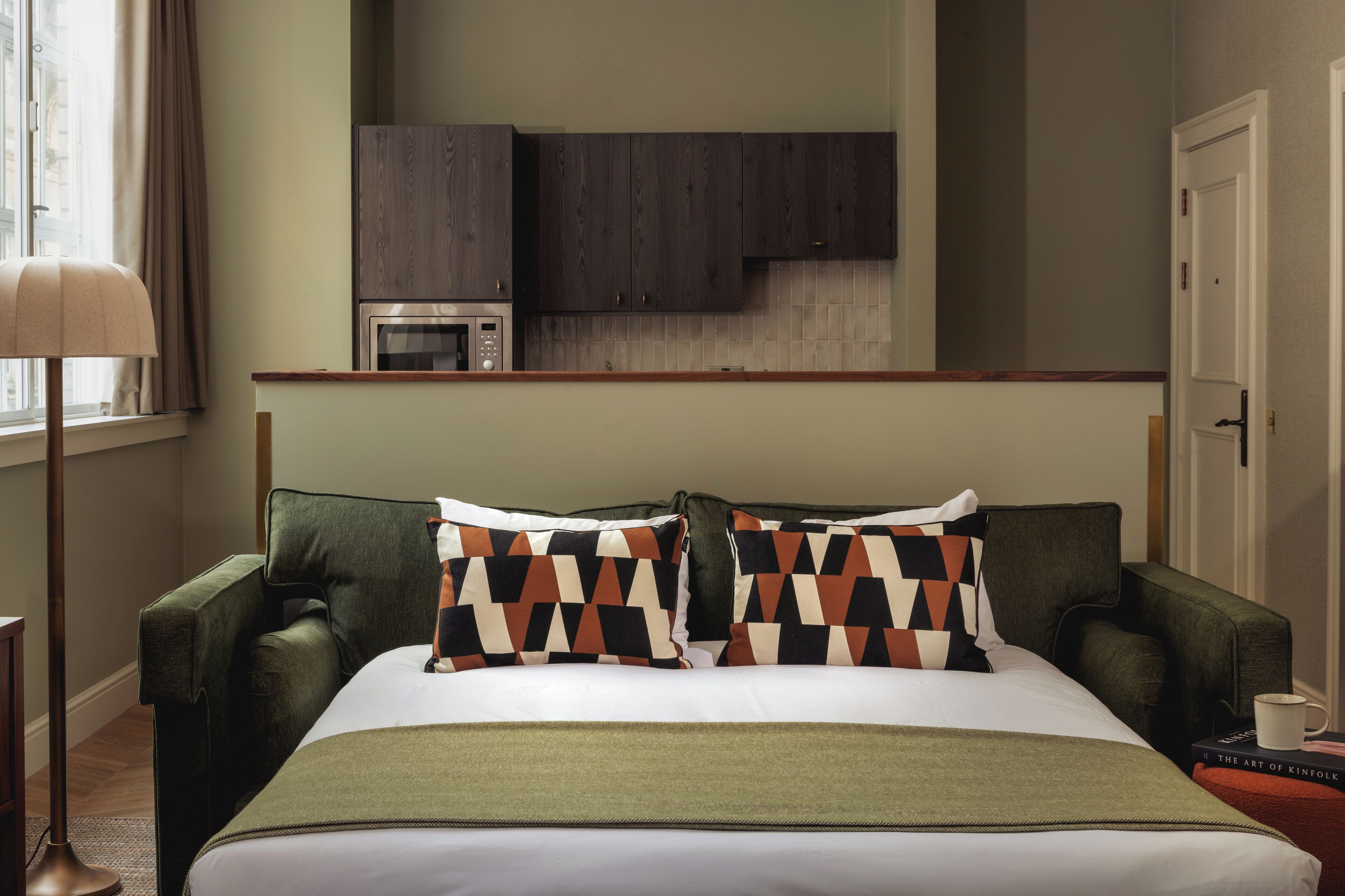 A hotel room with a bed, green armchair, and a kitchenette area.