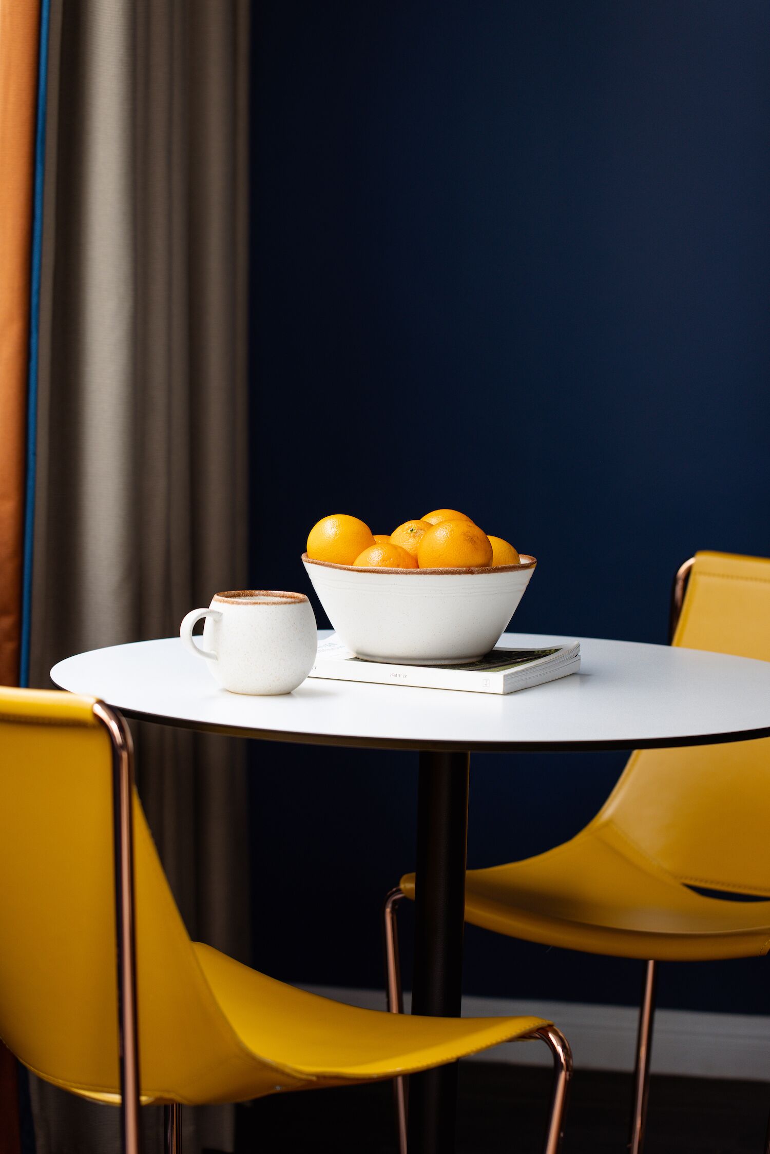 A round table with a white bowl of oranges and a white mug. Two yellow chairs are positioned around the table.