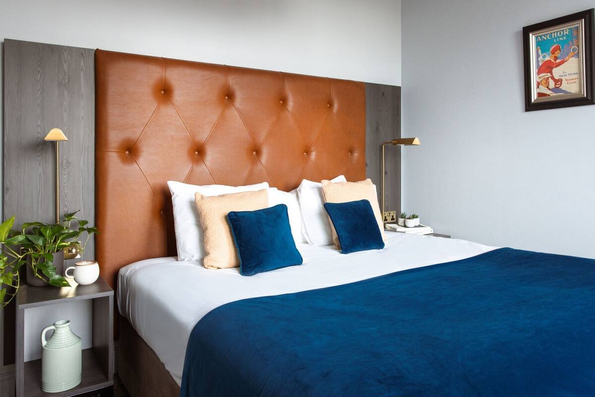 Quarto de hotel com cabeceira estofada, cama com roupa de cama branca e azul, mesa de cabeceira com abajur e um quadro na parede.