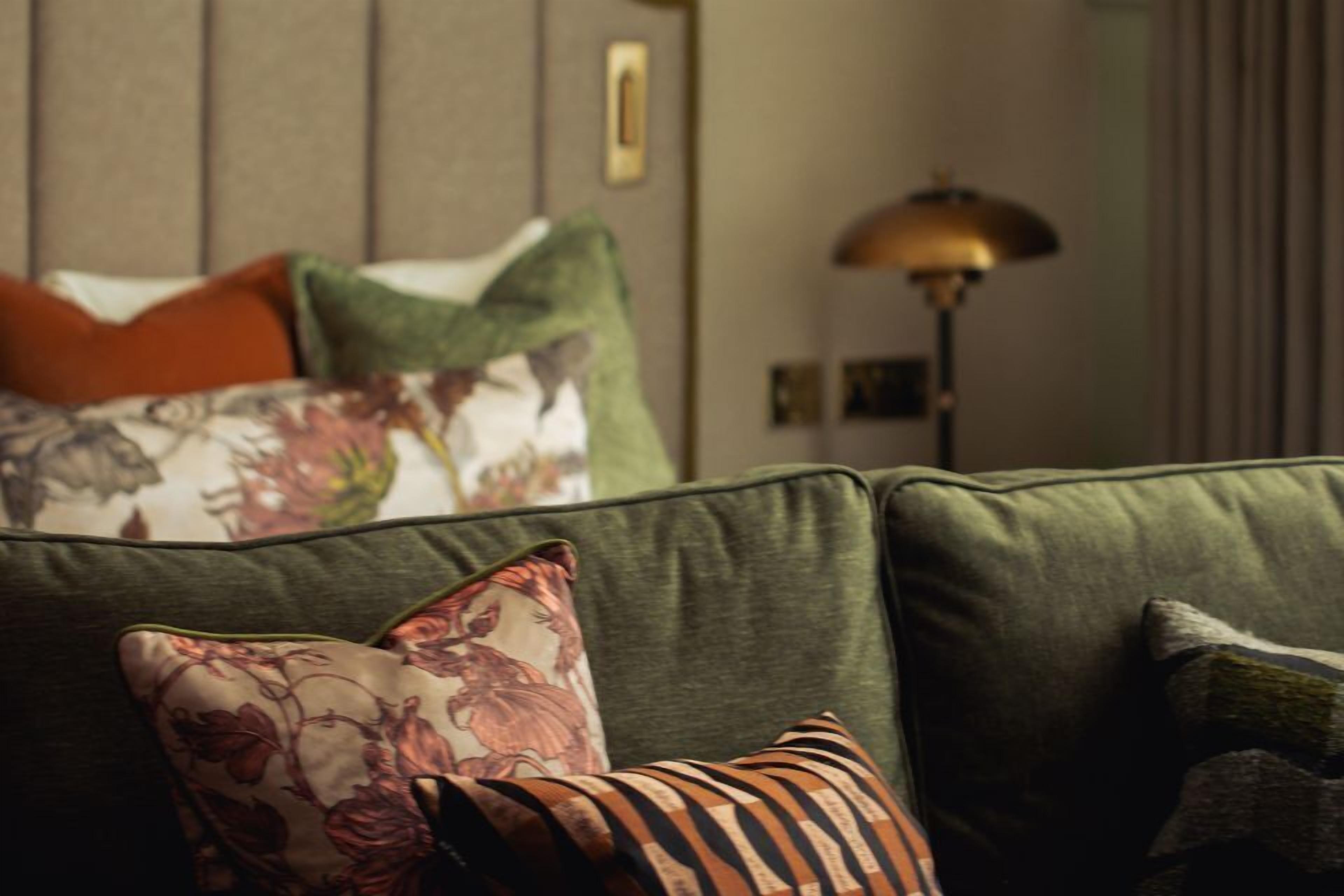Um sofá verde com almofadas estampadas de flores e listras, uma luminária e um quadro na parede.
