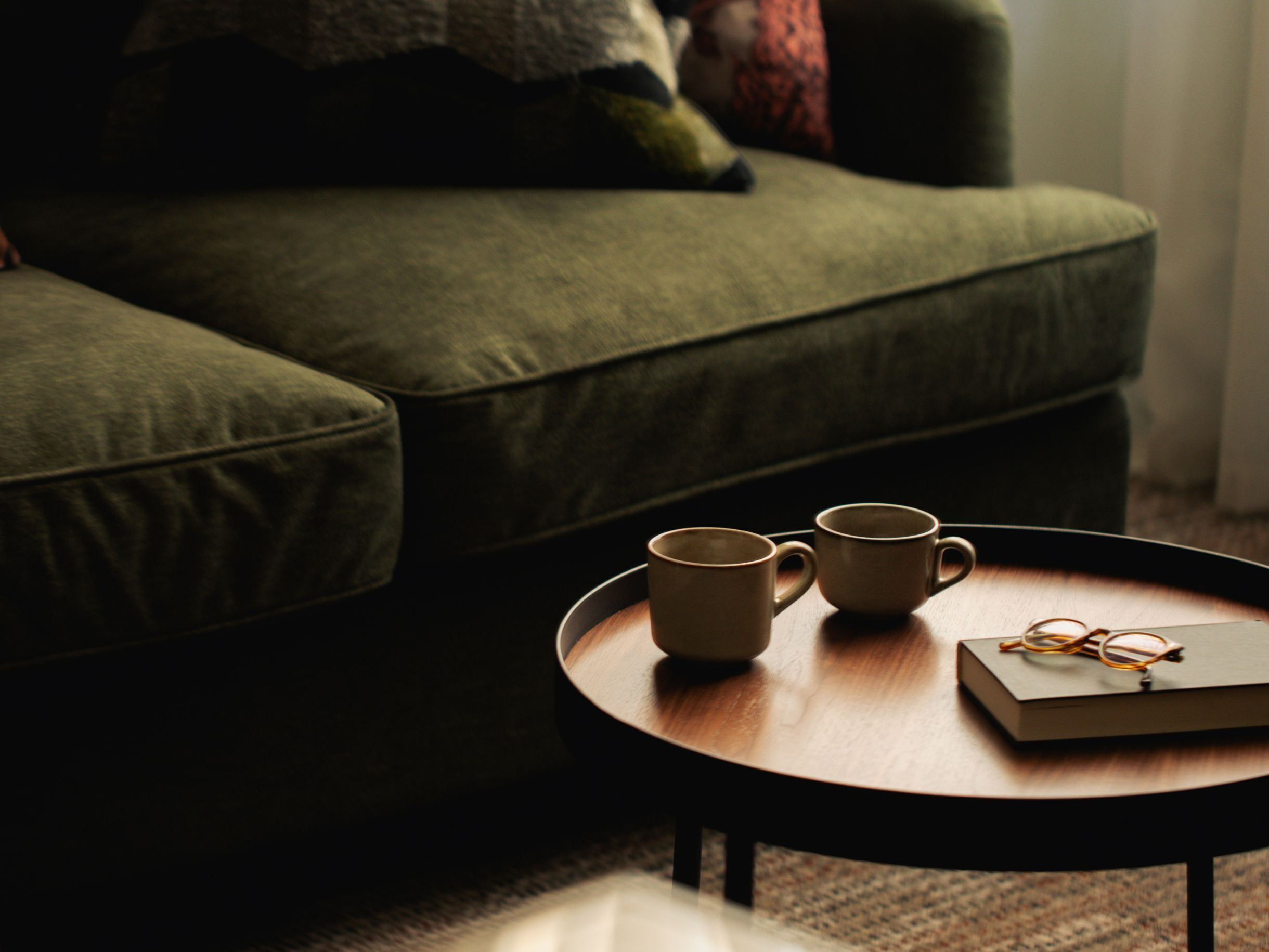 A green sofa with a pair of mugs and a book on a round wooden table.