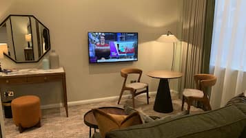 A hotel room with a TV displaying a news program, a desk with a mirror, two chairs, a small round table, a sofa, and a lamp.