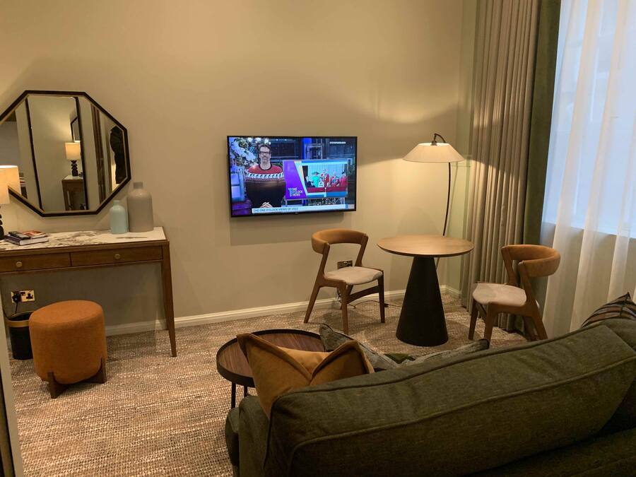 A hotel room with a TV displaying a news program, a desk with a mirror, two chairs, a small round table, a sofa, and a lamp.