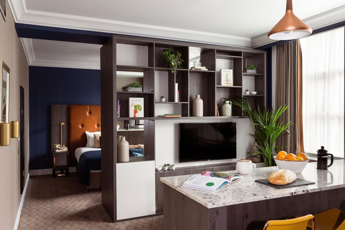 Quarto de hotel moderno com uma cama, uma televisão, área de refeições com cafeteira e uma estante de livros.