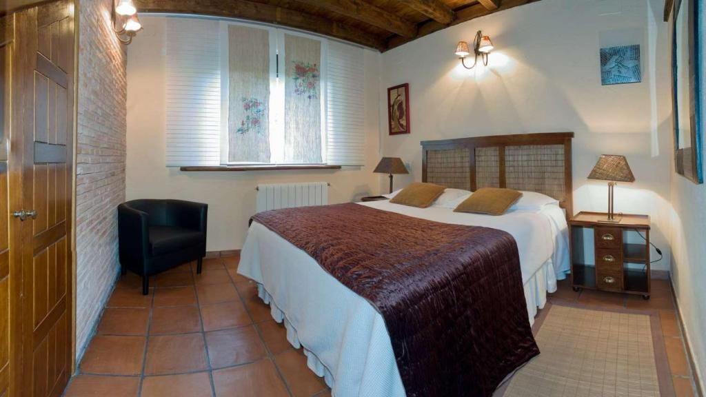 Un dormitori amb un llit gran, una cadira, una finestra amb persianes, un radiador i una làmpada penjada a la paret.