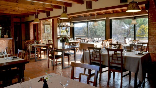 A restaurant with wooden beams, large windows, and multiple dining tables set with chairs and wine glasses. - SAIKA Rural (Puentes Viejas)