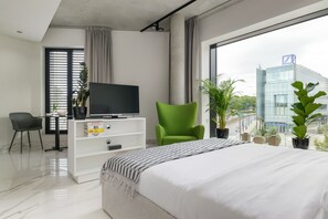 A modern hotel room with a large bed, a TV on a white cabinet, a green chair, and a view of a building through large windows.