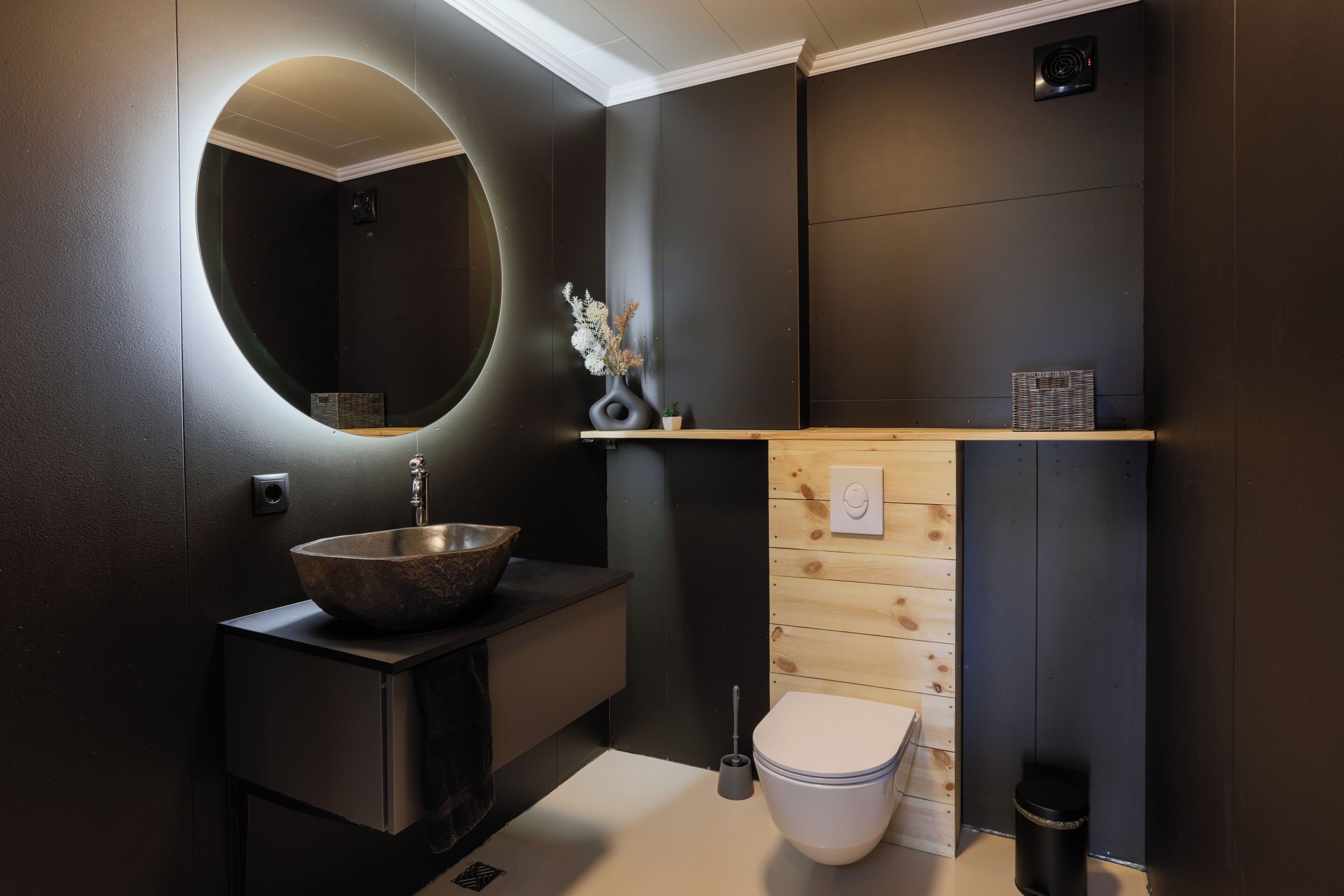 Un baño moderno con un espejo redondo, un lavamanos de pedestal, un inodoro y un pequeño estante con objetos decorativos.
