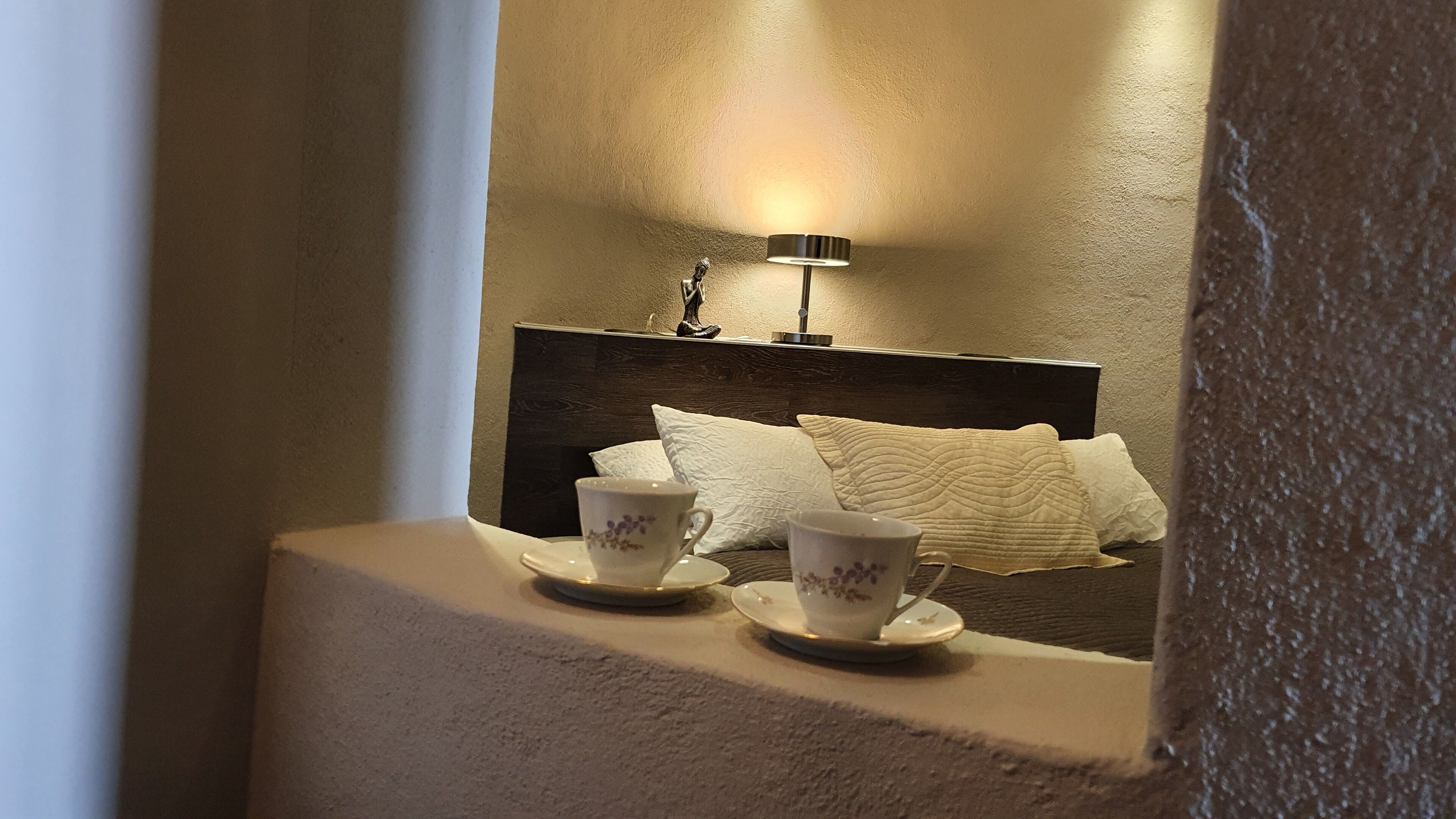 A bedroom with a bed, two cups on a bedside table, and a wall-mounted lamp.