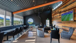 A modern lounge area with a long bench seating, a fireplace, and a large TV screen displaying a sports game. - Karuna Guesthouse (Skagafjörour)